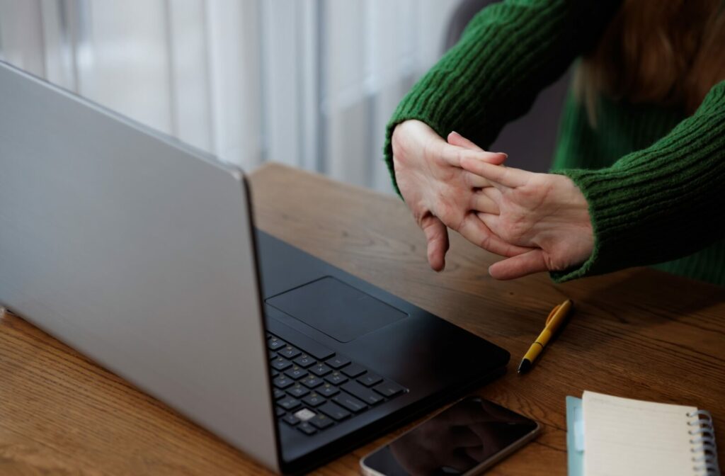 An adult in a green sweater stretching their hands while working at a laptop.